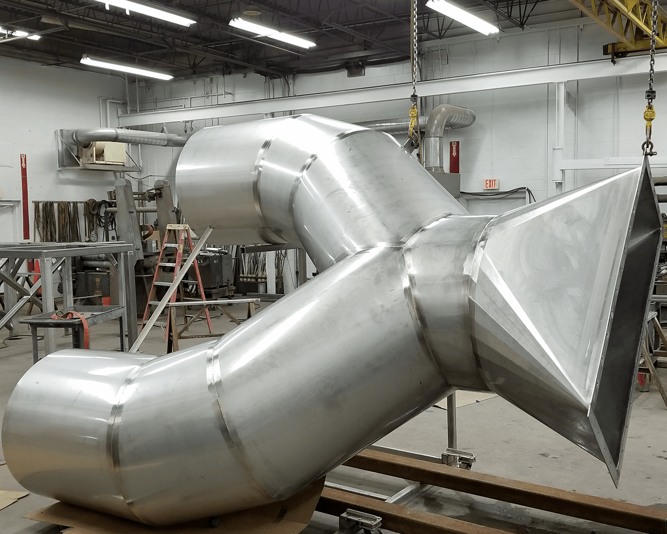 Prefabricated stainless steel HVAC duct transition assembly hanging in AET fabrication shop, ready for site delivery.